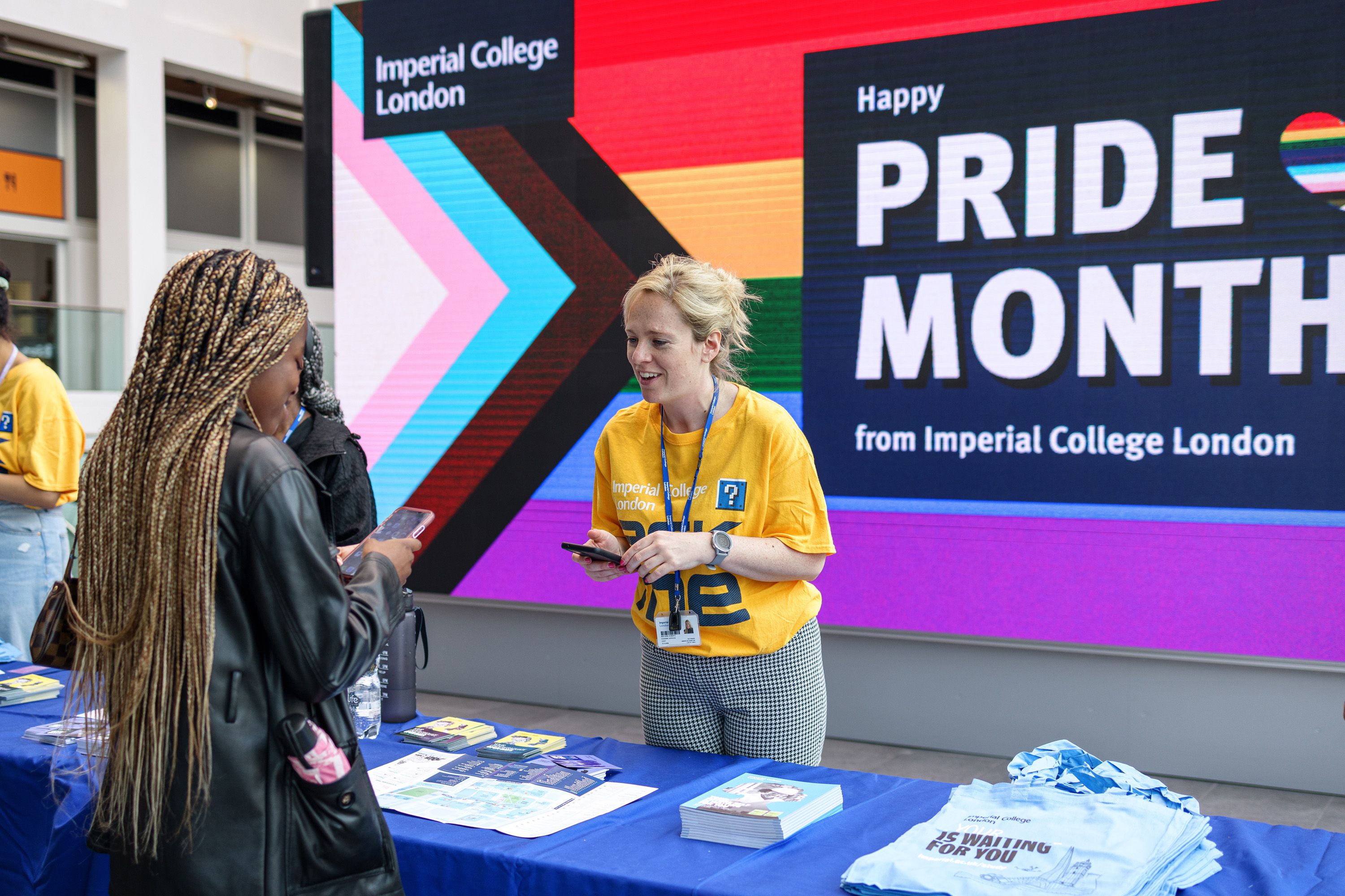 A woman in an yellow Ask Me T-shirt talks to a prospective student at a stand, with a sign behind them saying Happy Pride Month from Imperial College