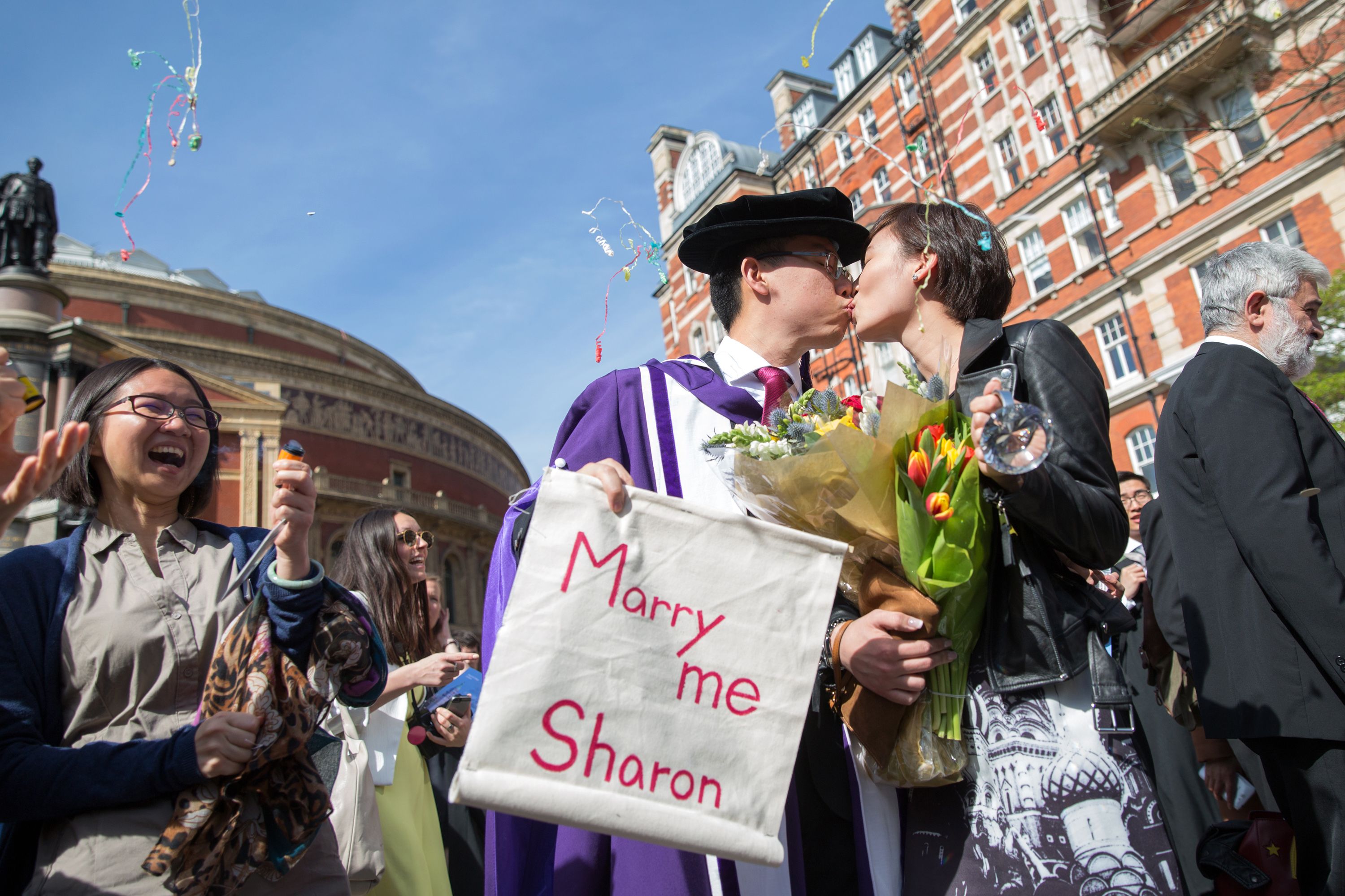 Two students in graduation robes kissing while the man holds a banner saying 