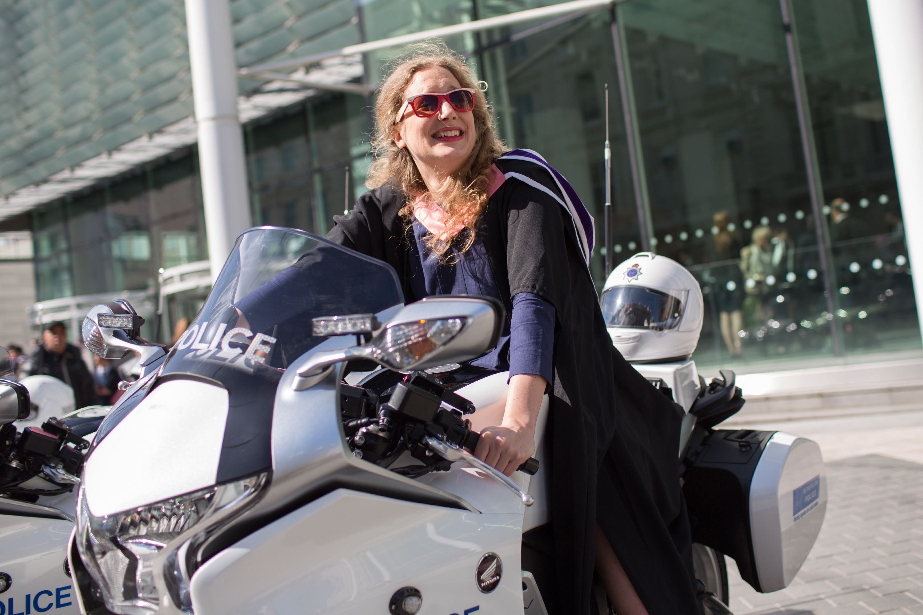 A female student in graduation robes and sunglasses sits on a police motorbike on Exhibition Road