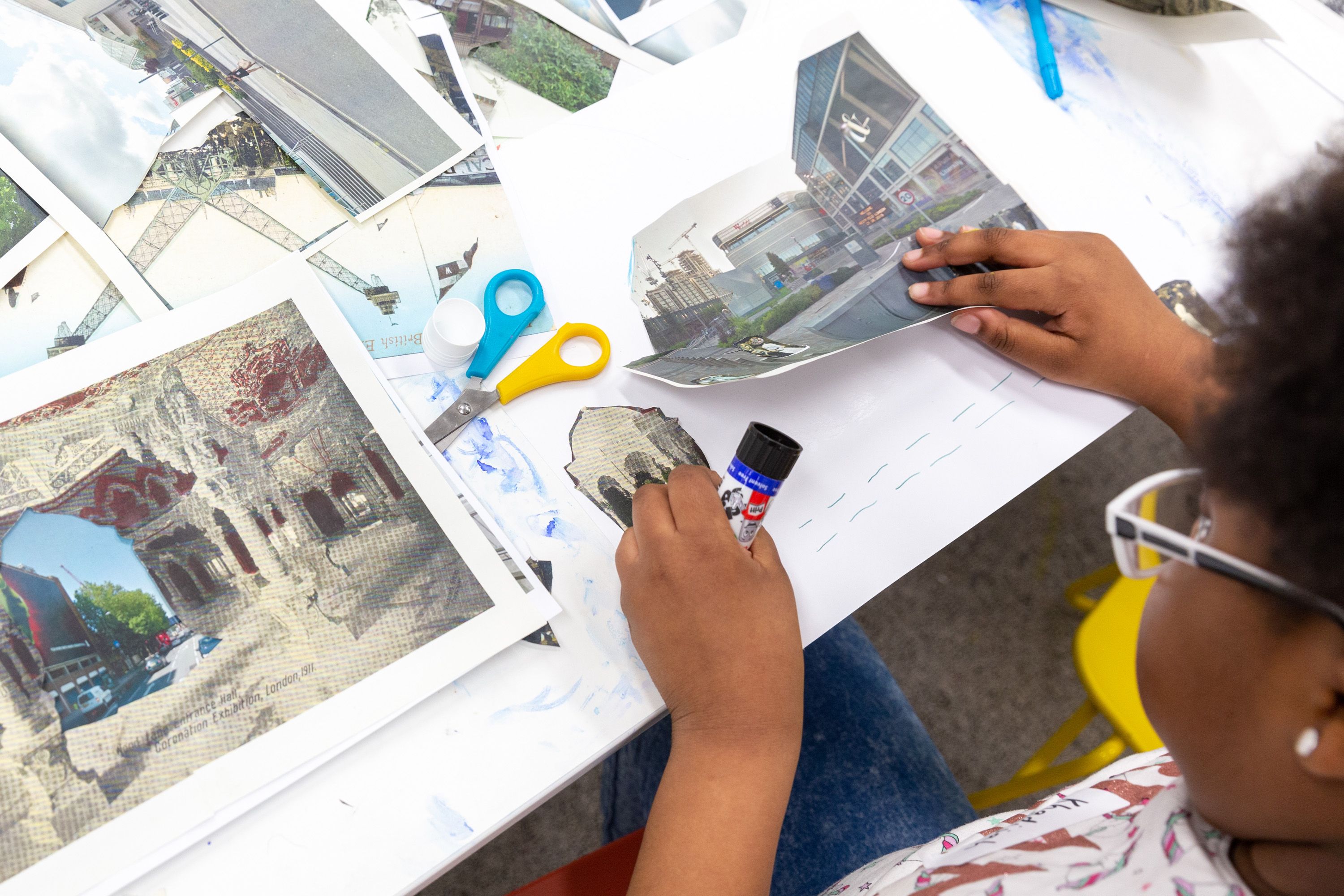 Seen from above, a child uses printed photos, pritt-stick glue and scissors to make a collage