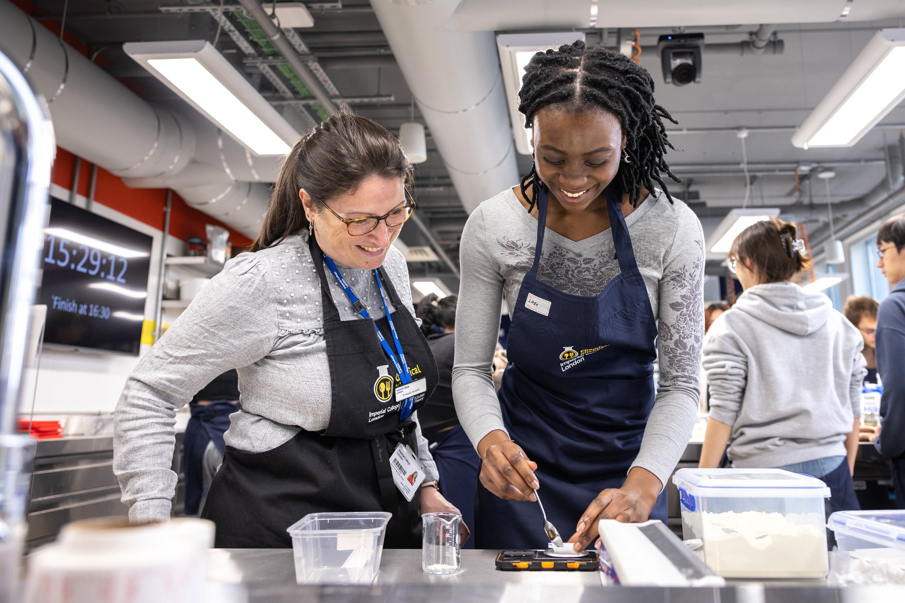 A female student and a supervisor smiling as they prepare materials on a metal work bench