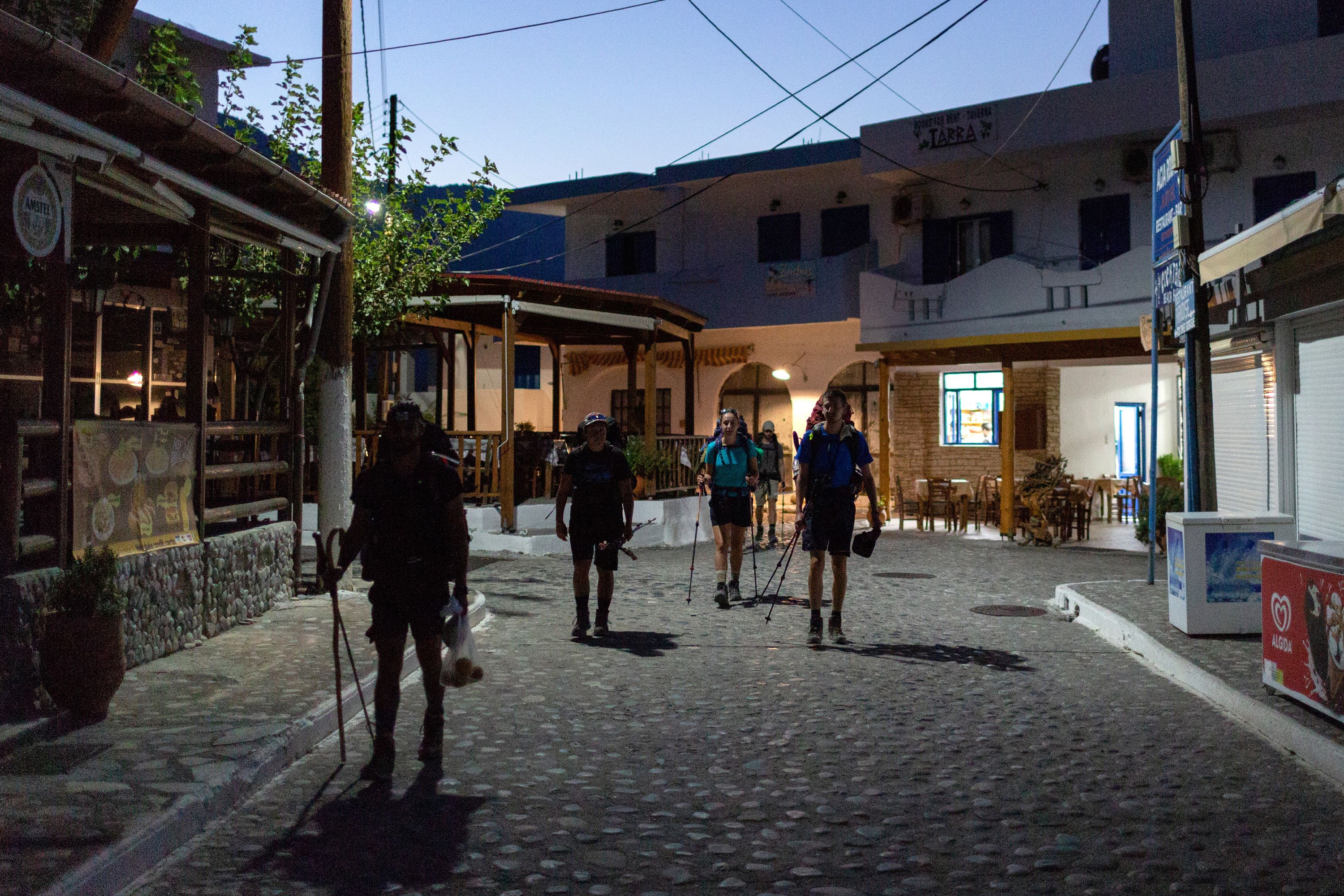 Students silhouetted as they walk through village streets at dusk, the streetlights casting long shadows on the cobbled street