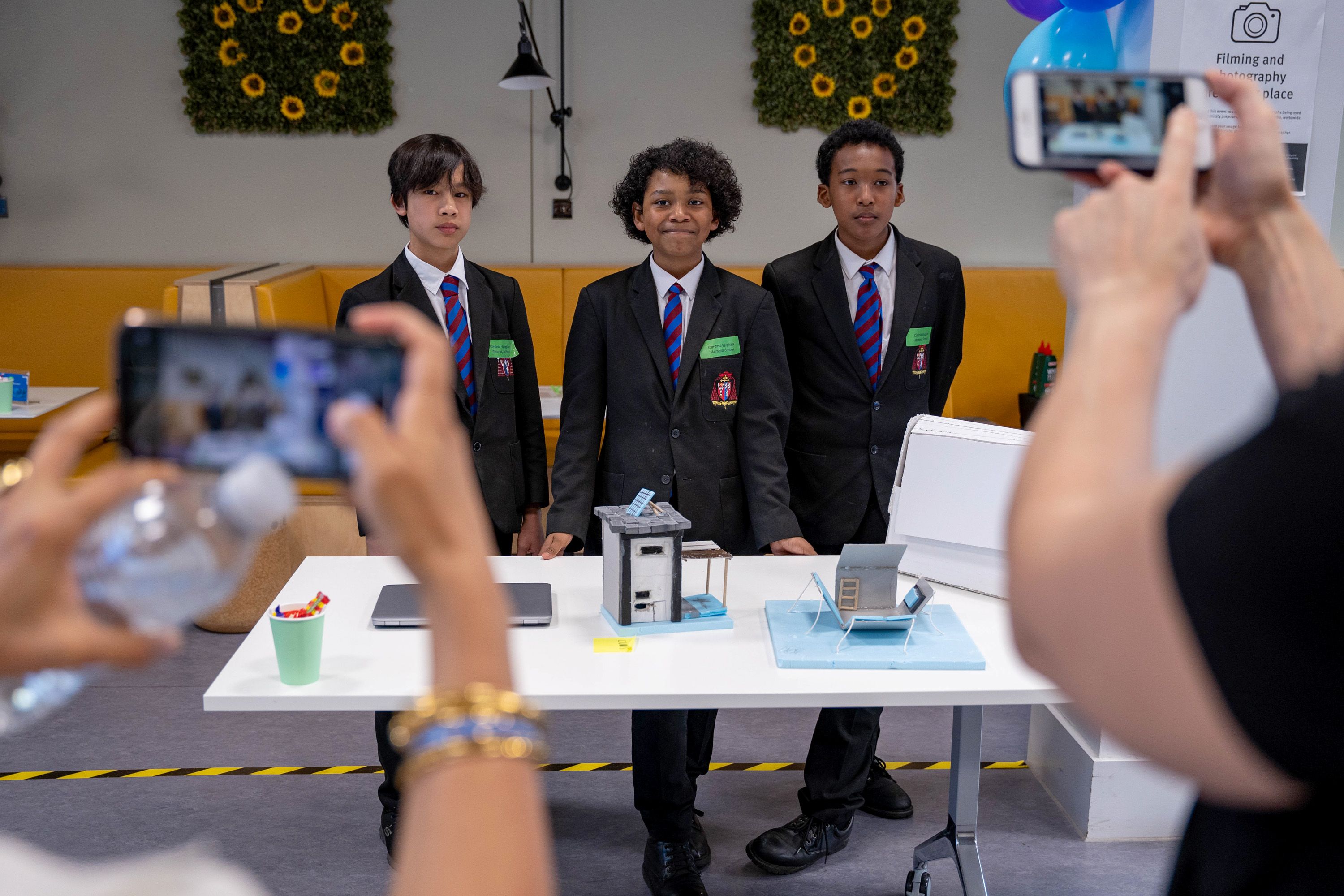 Three boys in school uniform stand proudly behind a table where there inventions are laid out, while in the foreground we see the phones of people taking photos of them