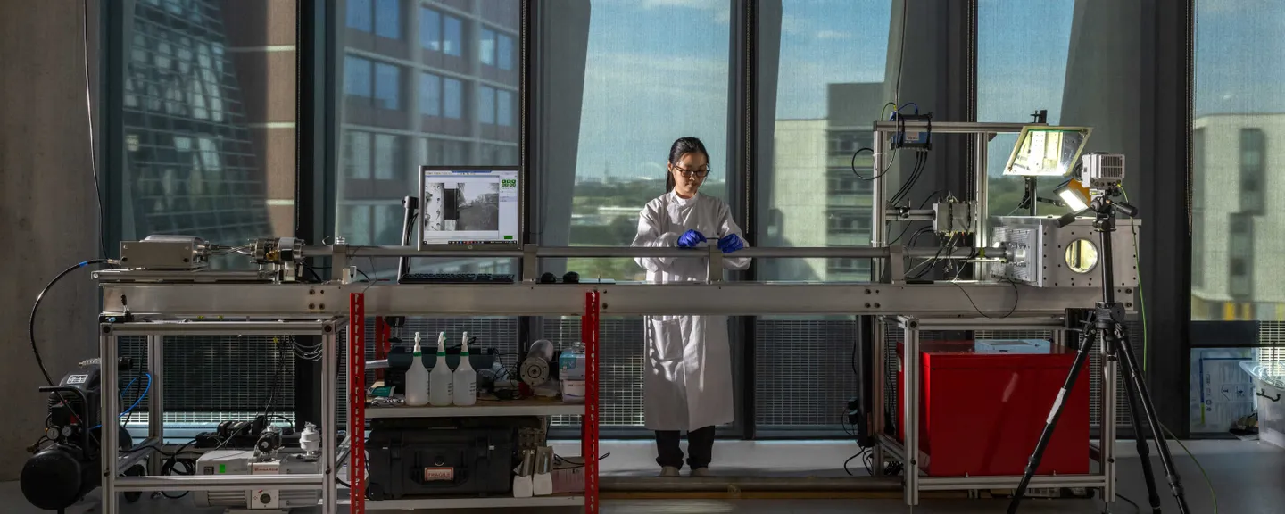 A researcher in a white lab coat and blue gloves works at a laboratory station with advanced equipment. The lab is bright, featuring large floor-to-ceiling windows with a view of modern buildings outside. The setup includes various instruments, a computer displaying a technical image, bottles, tools, and a tripod-mounted device.