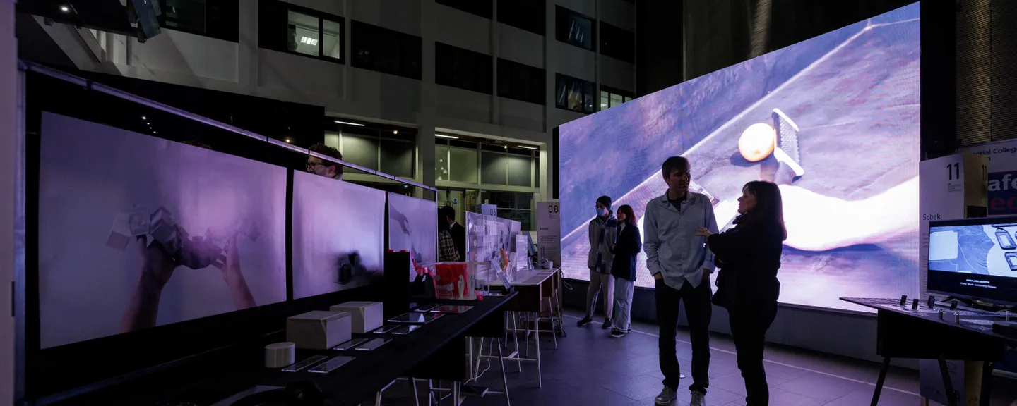 People talking in the main entrance of the college at the degree show private view for IDE and GID from the Dyson School of Design Engineering. A dark purple image lights up the space.