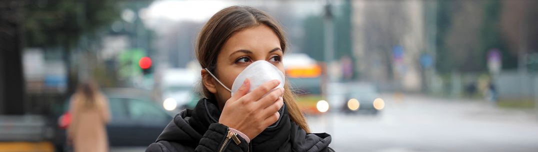 A woman in a black coat walking outside and wearing a white protective face mask