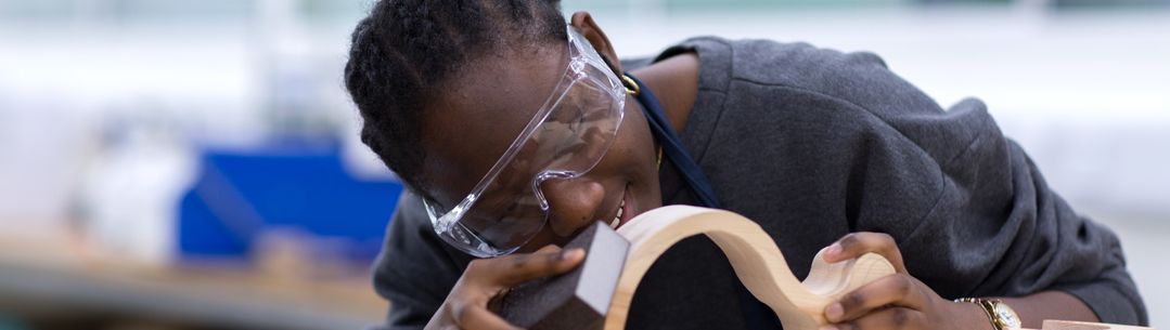 A young black woman wearing goggles and sanding a wooden object.