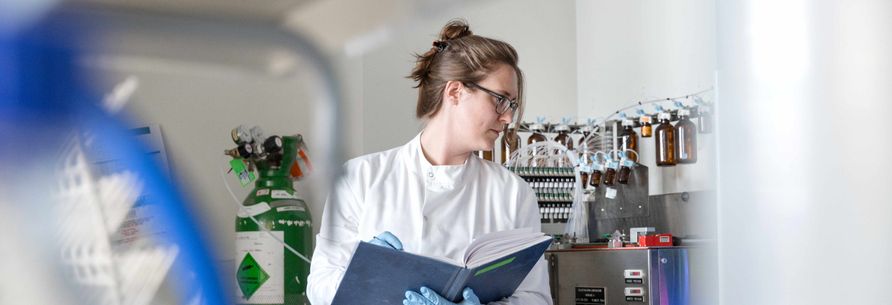 A woman is working in a lab, looking at an experiment and writing in a lab workbook.