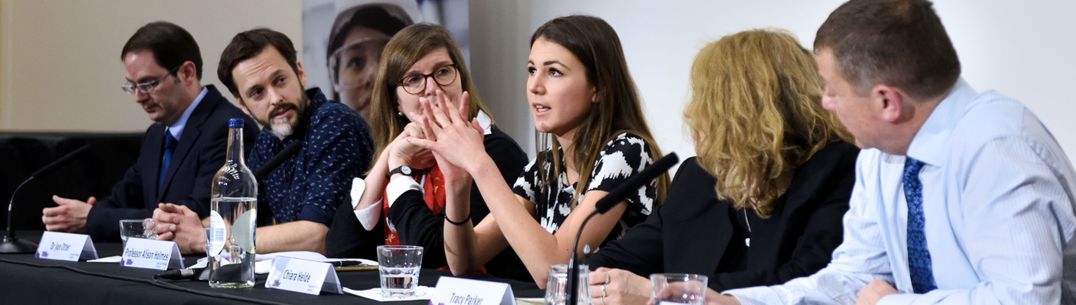 6 people on a panel at an event. A young woman in the centre is speaking, all other heads are turned towards her