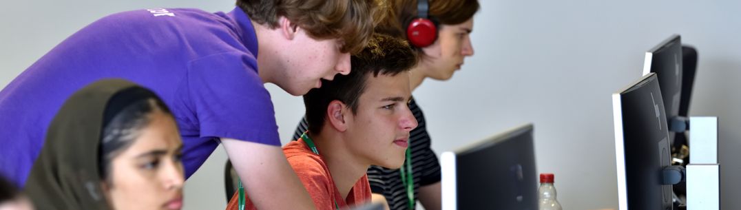 Three students sitting in a row looking at computer monitors with a mentor helping one of the students