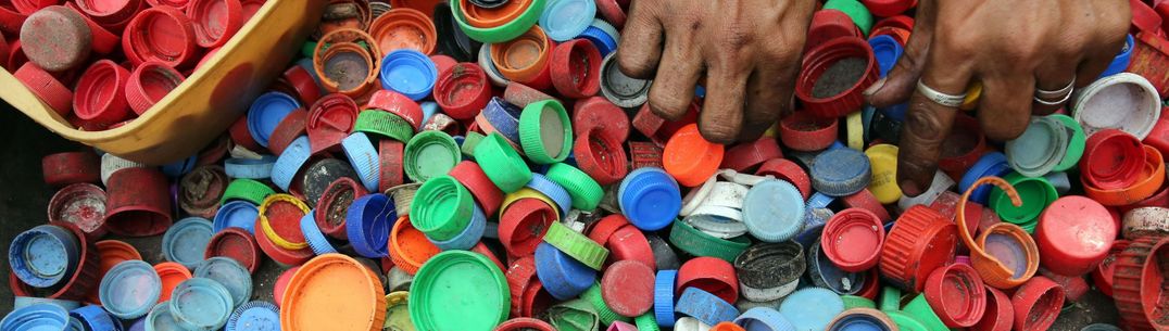 A large quantity of multicoloured plastic bottletops. A pair of adult hands is sorting red tops into a container