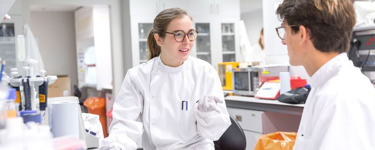 Two undergraduate students in lab coats talk at a lab bench