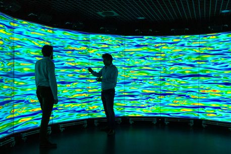 Two figures are silhouetted against a wall of screens showing a visualisation that looks like water rippling in blue, green and yellow