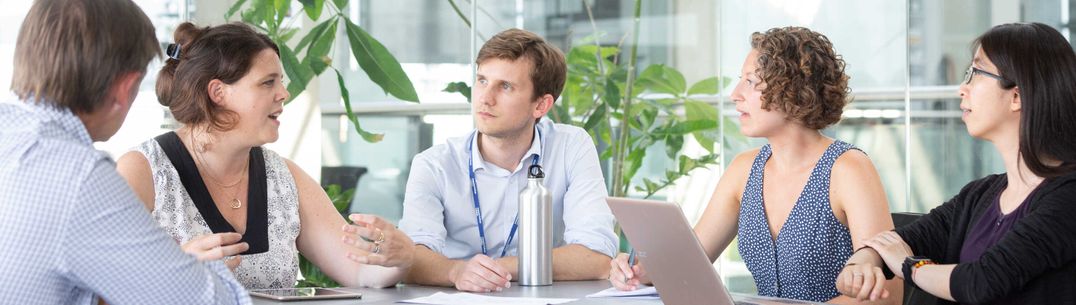 A group of staff in conversation in a glass meeting room with green plants