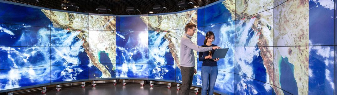 A man and woman look at a laptop together, in front of weather projections