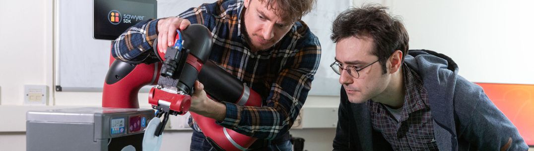 A researcher supports a robotic arm as another observes it unloading a mini-dishwasher