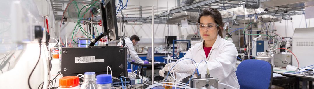 A researcher wearing a lab coat and safety glasses puts a solution in a bottle on a lab bench