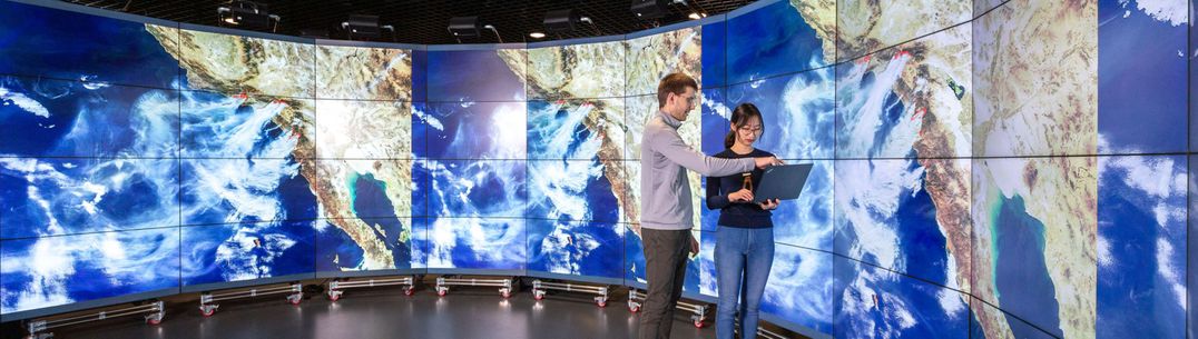 A student and an academic look at a laptop screen in the Data Observatory, with projections of satellite images of the Earth displayed behind them