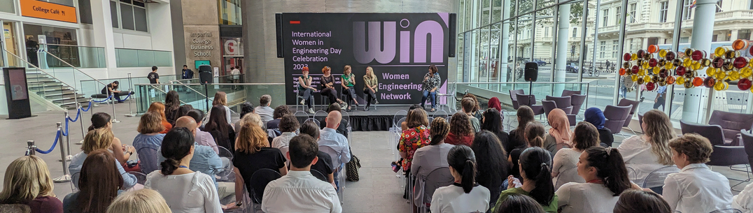 A panel of women on a stage in front of a screen, with audience in the foreground of the image.