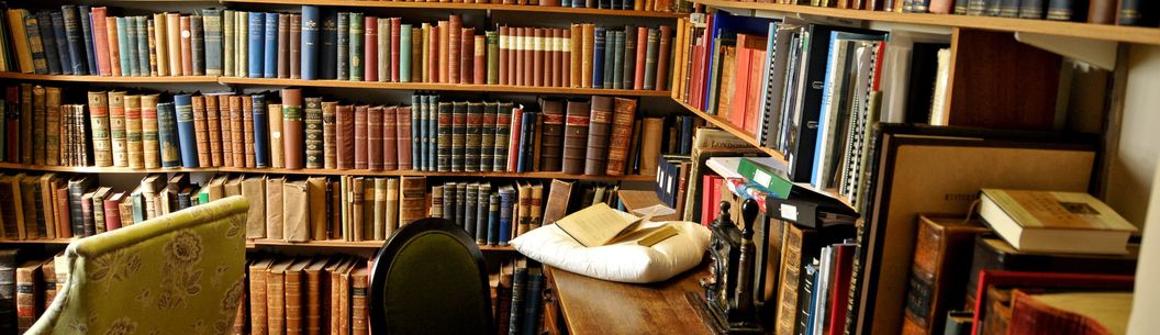 A library with walls covered floor to ceiling with old books, with a small desk and char tucked into the corner