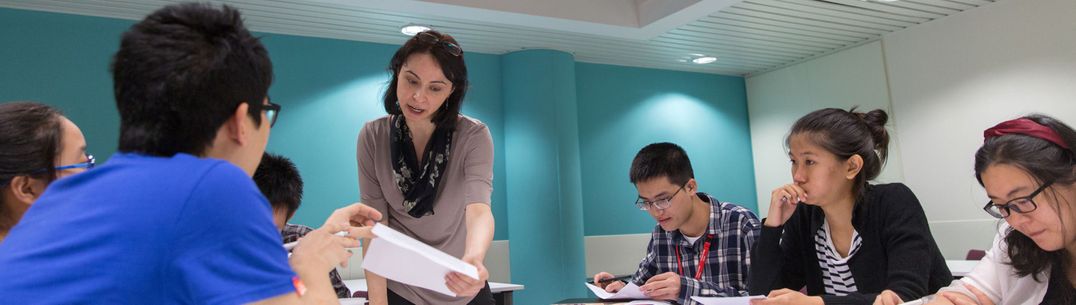 A group of students sitting around a desk in an academic English class talk to a teacher