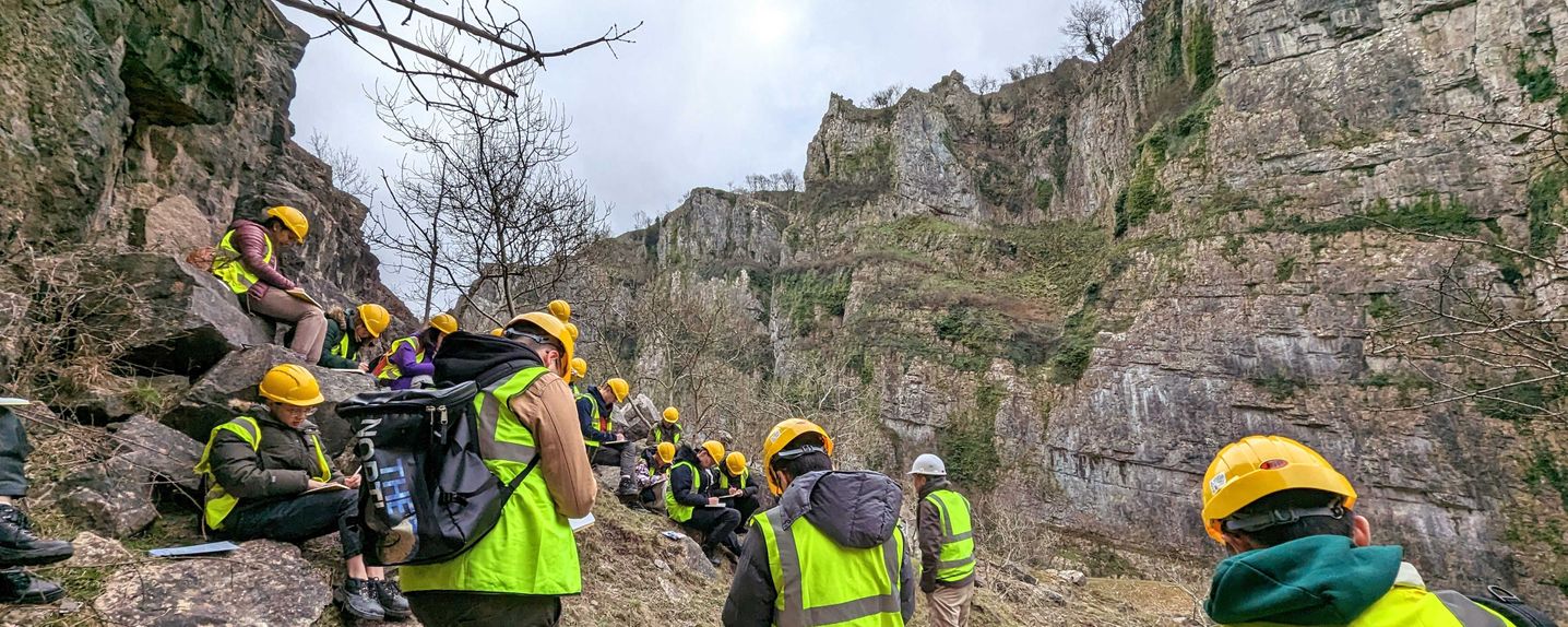 This image shows a group of 15 MSc students taking notes in high-vis jackets and hardhats. They are standing and sitting on a slope between the rocky faces of Cheddar Gorge investigating the bedding features.