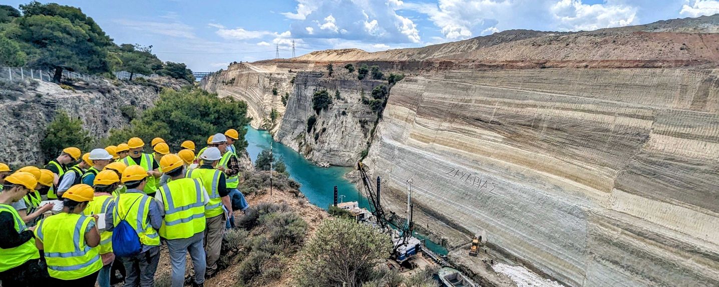 This image shows a group of roughly 20 MSc students and staff waring high-vis jackets and hard hats overlooking the Corinth Canal. They take notes looking down at the water and excavations taking place below.