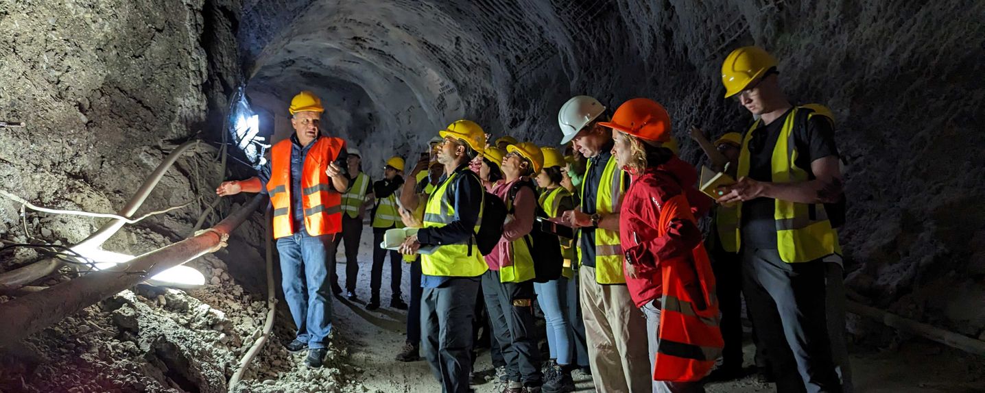 This image shows a group of roughly 20 MSc students and staff waring high-vis jackets and hard hats in an underground tunnel. They are being taught about the structure and excavation from a Greek engineer standing in the light.