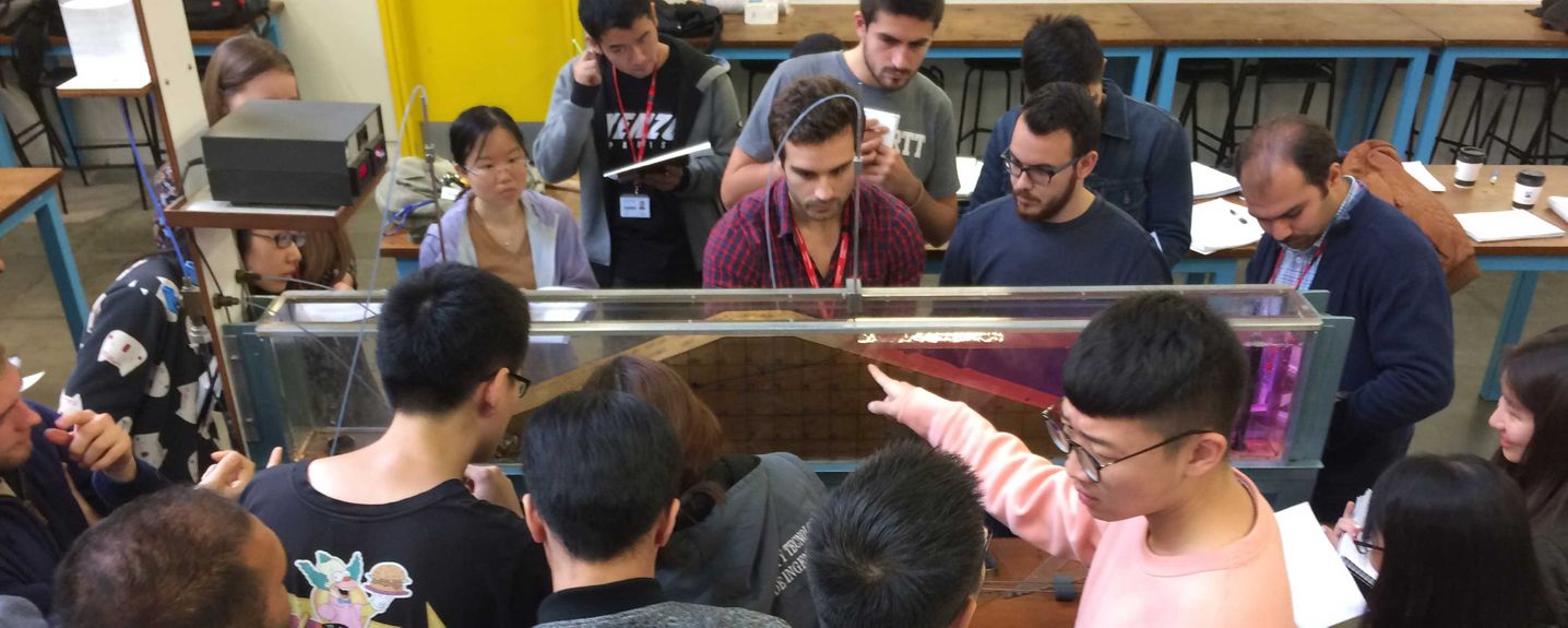 This image shows a group of circa 15 MSc students congregating around a model dam.  They are discussing the flow patterns they can see.  One student is taking a measurement.  All are pretty focussed on the activity.  There are about 12 male students and 4 female students.  The image was taken in the flexible teaching space for laboratory work.