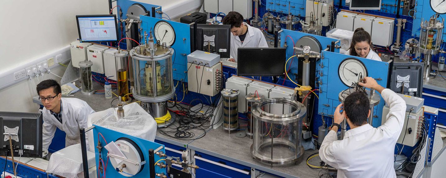 This image is a view of the geotechnical research laboratory taken from a mezzanine level.  It shows 4 researchers (3 male, 1 female) standing at computers adjacent to laboratory apparatus.  Three triaxial cells are visible.  Much of the imaging shows tubing, connections and pressure controllers.