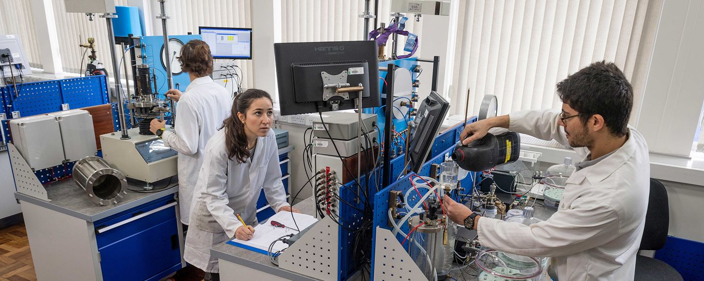 This image shows three PhD students (2 male, 1 female) in a research lab.  3 lab benches are visible.  The only piece of equipment with a clear view is a triaxial cell.  Computer screens that show data from the equipment are visible.