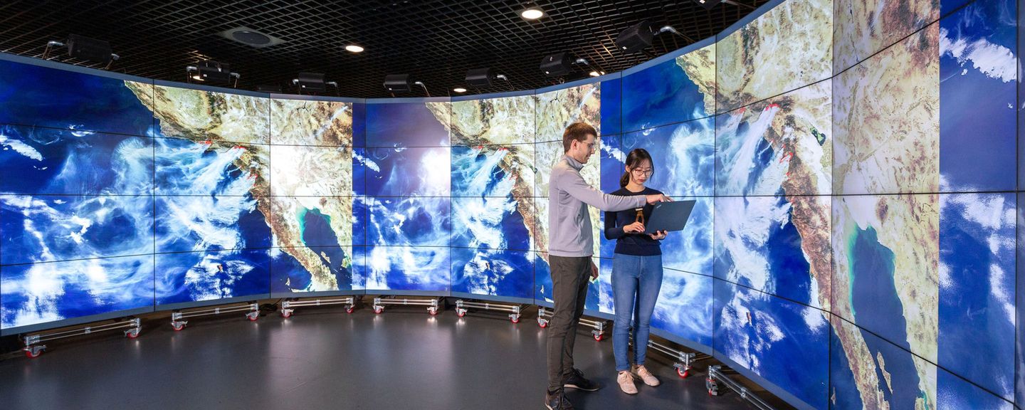 A student and an academic look at a laptop screen in the Data Observatory, with projections of satellite images of the Earth displayed behind them