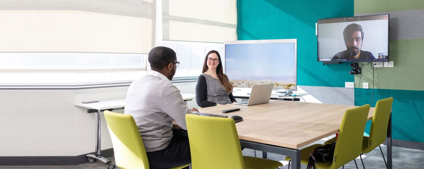 two people sit at a meeting table with another person joining the meeting via a large screen