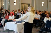 Woman stands at the front of conference room giving a talk and gesturing to presentation on screen