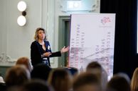 Woman delivering talk stands in front of conference hall and gestures to screen