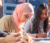 Two women sit at table outside in the sun and fill out form