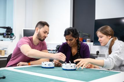 Three students test switches on small robotic devices on a table top