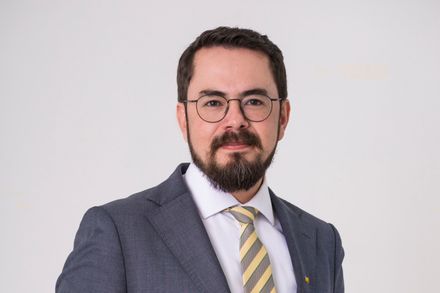 Headshot of Dr Inty Grønneberg - he is wearing a yellow striped tie and grey blazer, standing against a white-grey backdrop