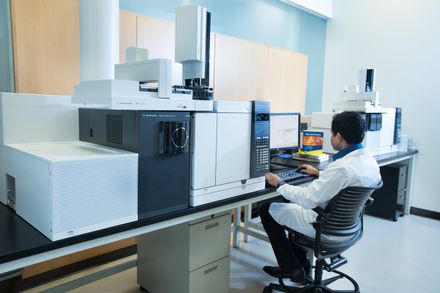 Person in a lab coat sitting at a computer attached to scientific instrument