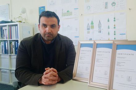 Ummer in his office at Empace, he is wearing a brown blazer over a black t shirt and sits at a desk with his hands joined together in front of some certificates