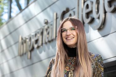 A student with long hair and glasses smiles as she stands beneath a sign for Imperial College London