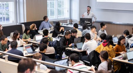 A teacher standing in a busy classroom while students work on laptops at group tables