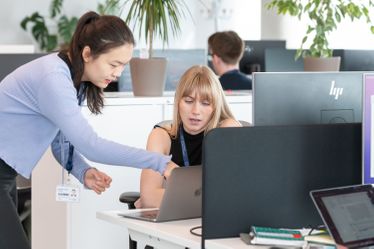 Early career researchers talking at their desks and one leans over to point at others laptop screen