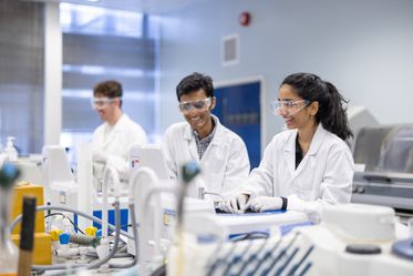 Three undergraduate Materials students sit in the lab. They are wearing safety goggles, gloves and lab coats and smile.