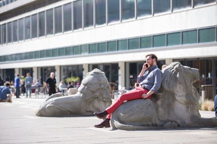 A student talks on his mobile phone while sitting on one of the lion statues in front of the Central Library and Dangoor Plaza at the South Kensington Campus