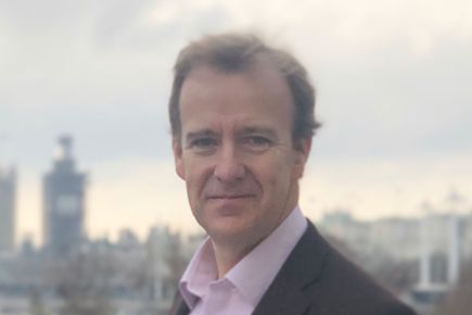 Portrait of Oliver Woolley looking at the camera, in the background is a cityscape with a stormy sky