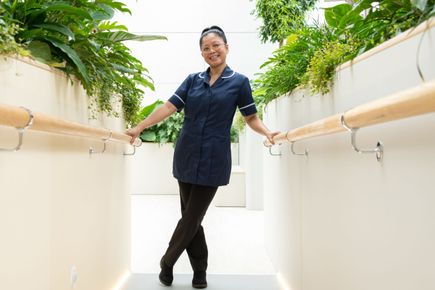 A woman in scrubs leans on bannisters in a corridor with plants