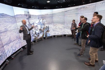 A man talks to a crowd in front of a wide screen showing an image of the Mars rover
