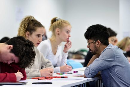 A tutor kneels down to speak to a student in a workshop. Two other students continue with their work.