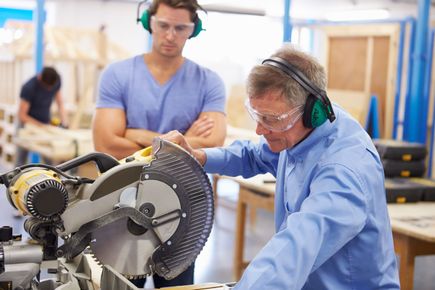 A workshop operative using a circular saw wearing ear defenders and protective glasses whilst another operative watches on